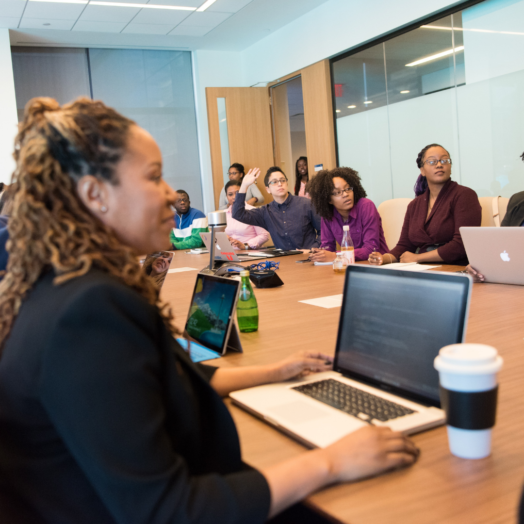 A group of people of different races, but predominately Black women are sitting around a table. Many of the people have their laptops open in front of them. Their attention is to the front of the room, and one person has their hand raised.