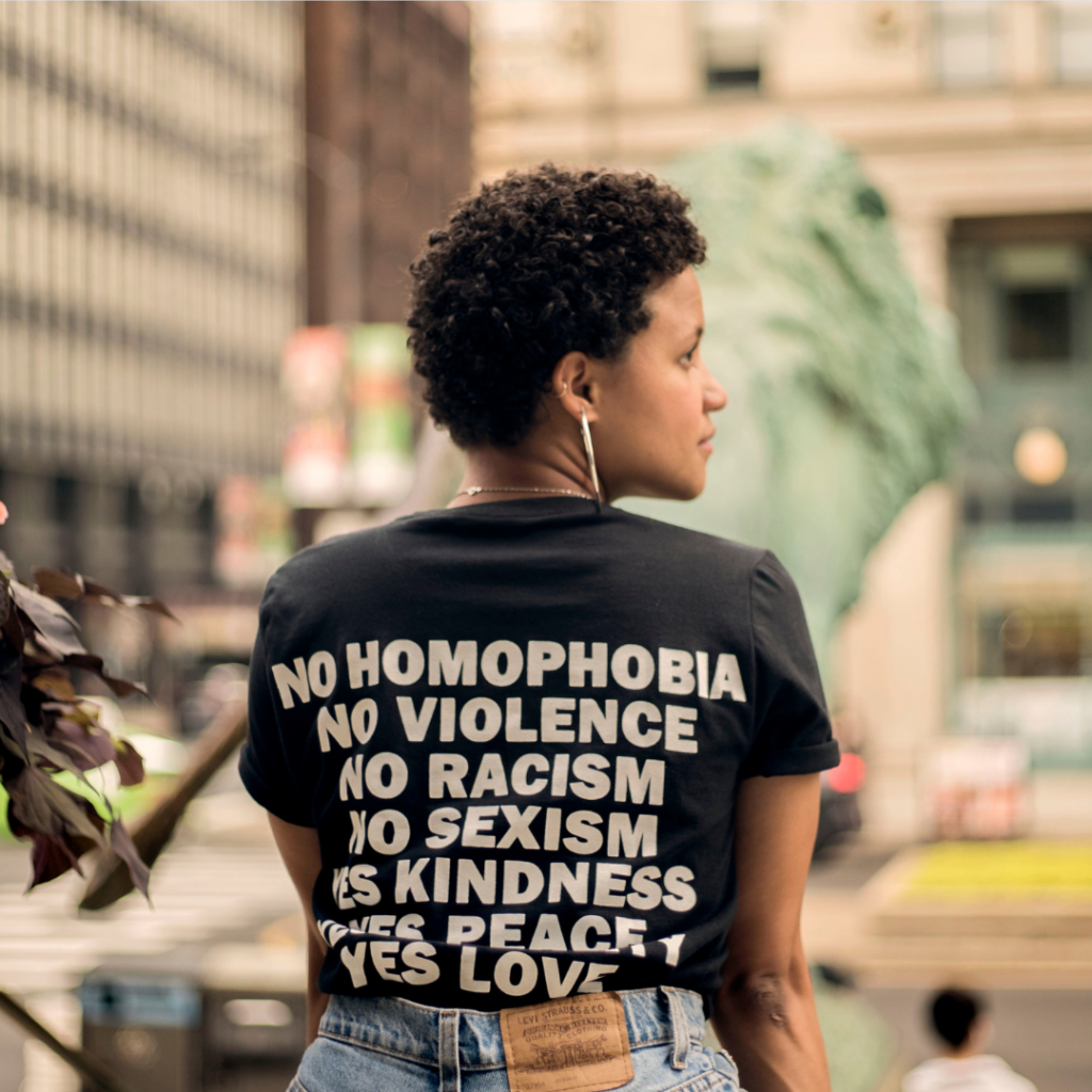 Black woman with short natural hair wearing a t-shirt that says "No Homophobia, No Violence, No Racism, It's Kindness, Yes to Love"