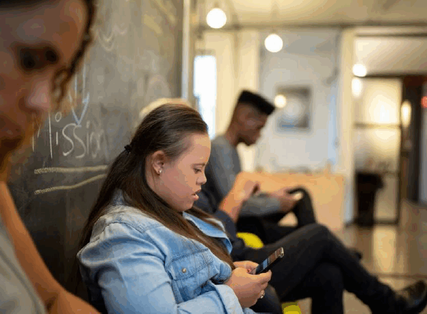 A young white woman with a developmental disability sits by a blackboard, reading on her phone.